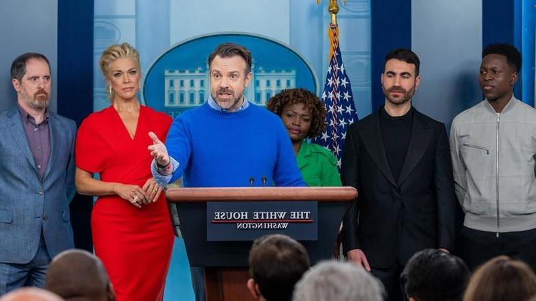 Six people stand behind a podium bearing a sign that reads "The White House"